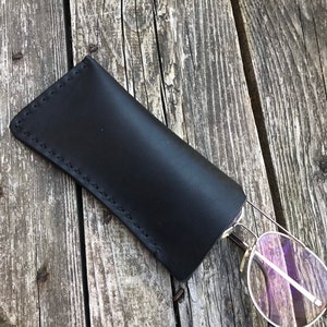 May include: Black leather glasses case with a stitched edge. The case is lying on a wooden surface with a pair of glasses sticking out.