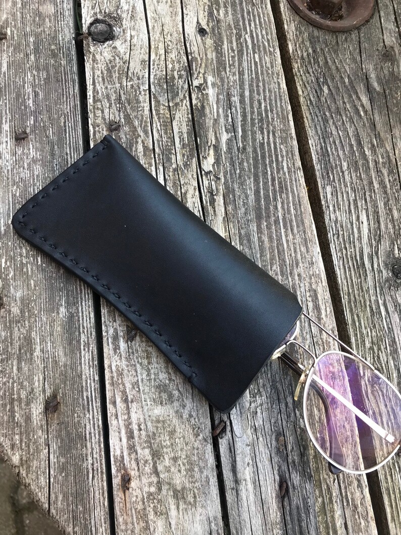 May include: Black leather glasses case with a stitched edge. The case is lying on a wooden surface with a pair of glasses sticking out.