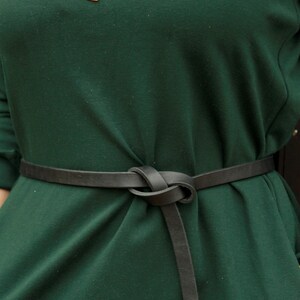 May include: A dark green dress with a black leather belt tied in a knot at the front. The belt is worn high on the waist and has a long strap that hangs down the front of the dress.