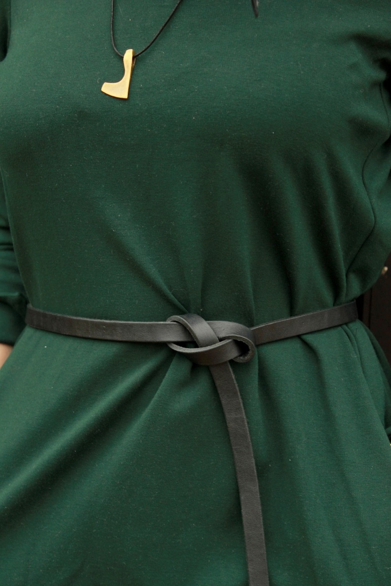 May include: A dark green dress with a black leather belt tied in a knot at the front. The belt is worn high on the waist and has a long strap that hangs down the front of the dress.