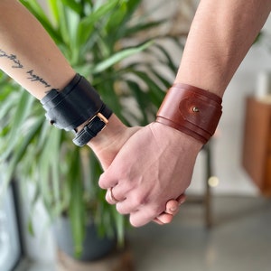 Pode incluir: Duas pessoas de mãos dadas, cada uma usando um bracelete de couro. O bracelete da esquerda é preto e o bracelete da direita é castanho.