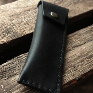 May include: A black leather pencil case with a flap closure and a silver button. The case is stitched with white thread.