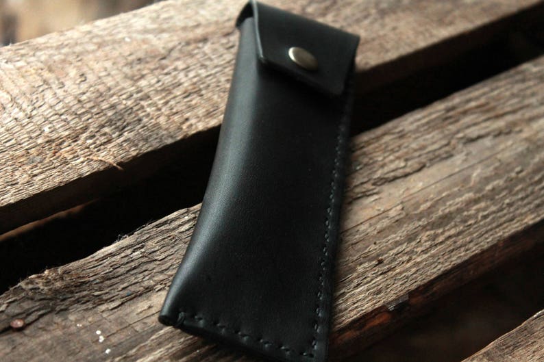 May include: A black leather pencil case with a flap closure and a silver button. The case is stitched with white thread.