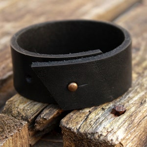 May include: A black leather cuff bracelet with a triangular flap and a small copper rivet.