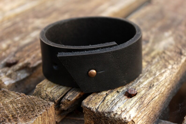 May include: A black leather cuff bracelet with a triangular flap and a small copper rivet.