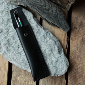 May include: A black leather pencil case with two pencils inside. The case is lying on a gray rock.