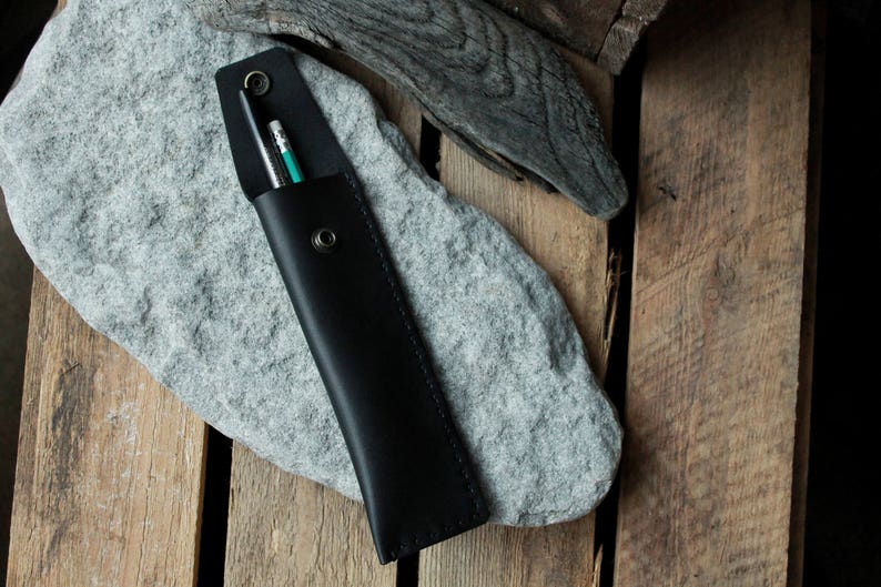 May include: A black leather pencil case with two pencils inside. The case is lying on a gray rock.