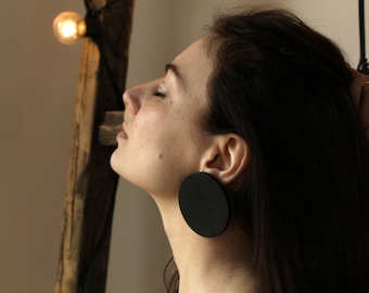 May include: A woman with long dark hair wearing large black round earrings. The earrings are made of a smooth, matte material.
