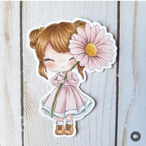 Puede incluir: Una pegatina con una niña de dibujos animados con un vestido rosa que sostiene una gran margarita rosa. La niña tiene el pelo castaño recogido en moños y lleva botas marrones. La margarita tiene un centro amarillo y un tallo verde.