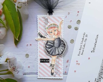 May include: A pink and black ballerina bookmark with a black tutu, a pink flower crown, and the word "DREAM" printed on the bookmark. The bookmark is decorated with pink and white stripes and black polka dots.
