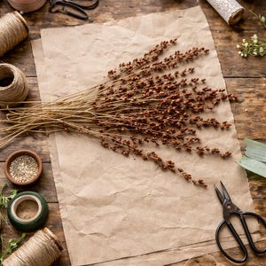 May include: A rustic still life arrangement featuring craft supplies on a wooden surface. Brown twine, pink ribbon, scissors, and dried flowers are arranged around a sheet of brown paper. The scene evokes a sense of crafting and natural elements.