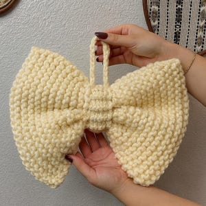 May include: A large, cream-colored, hand-knitted bow-tie shaped wall hanging. The bow has a loop at the top for hanging. The yarn is thick and the stitches are visible. The bow is held by two hands.