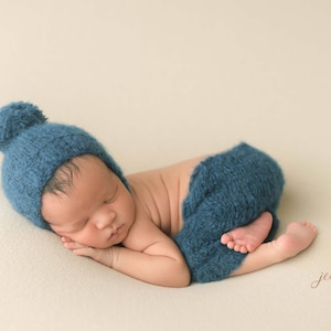 May include: A newborn baby wearing a blue knitted hat with a pom-pom and matching blue knitted pants, sleeping on a cream-colored background.