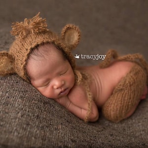 Puede incluir: Un bebé recién nacido con un gorro de punto marrón con orejas y un conjunto de punto a juego. El bebé está durmiendo sobre una superficie marrón texturizada.