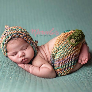 May include: A sleeping baby wearing a multi-colored knitted hat and matching pants with a button. The baby is lying on a green textured surface.