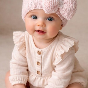 May include: A baby wearing a light pink knitted hat with a large bow and a light beige romper with wooden buttons. The baby has blue eyes and is smiling.
