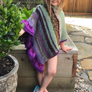 Può includere: Un poncho colorato lavorato a mano a maglia nei toni del viola, verde e marrone. Il poncho presenta un collo alto e un bordo con frange. La bambina indossa infradito blu.