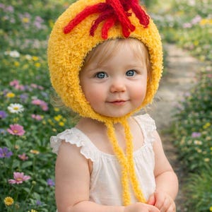 Könnte beinhalten: Ein Kind trägt eine gelbe, flauschige Mütze mit roten Akzenten und langen gelben Bändern. Das Kind trägt ein weißes Kleid und steht in einem Garten voller bunter Blumen.