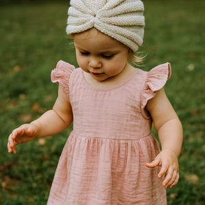 Puede incluir: Una niña pequeña con un vestido rosa claro con mangas con volantes y un turbante de punto color crema. El vestido tiene cintura fruncida y cuello sencillo. La niña está de pie sobre hierba.