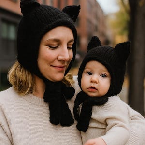 以下が含まれることがあります： お母さんと赤ちゃんが、お揃いの黒いニットの猫耳帽子をかぶっています。帽子には尖った耳があり、お揃いのスカーフと組み合わせています。どちらも明るい色のセーターを着ており、居心地の良いコーディネートされた外観になっています。
