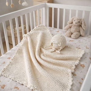 May include: A cream-colored knitted baby blanket and bonnet set with a scalloped edge, resting on a crib mattress with a subtle animal print. A stuffed bunny and other nursery decor are visible in the background.