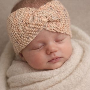 May include: A newborn baby wearing a hand-knitted headband in shades of beige, peach, and blue. The baby is wrapped in a soft, cream-colored blanket. The headband has a knotted design.
