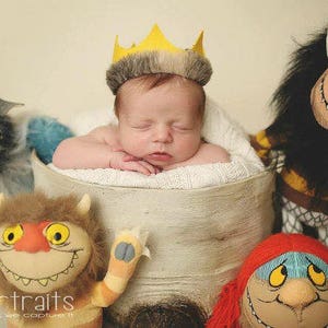 May include: A newborn baby wearing a yellow crown sleeps in a white basket surrounded by four plush toys. The toys are a blue and gray bull, a red and blue monster, a brown and yellow monster, and a brown and yellow monster with a red mane.