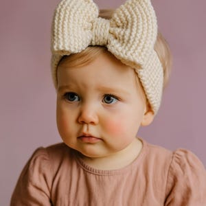 May include: A cream-colored knitted headband with a large bow detail. The headband is worn by a baby with fair skin and blue eyes, wearing a pink dress. The background is a solid purple color.