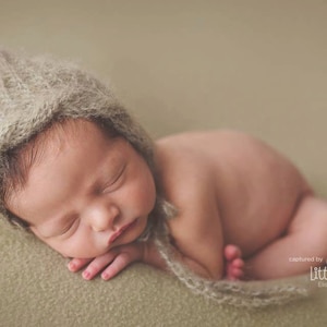 Op de afbeelding: Een pasgeboren baby die een lichtbruine gebreide muts draagt, slaapt op een lichtbruin, getextureerd oppervlak. De baby is opgerold met de armen onder de kin. De muts heeft een lange, stromende stropdas die over het lichaam van de baby is gedrapeerd.
