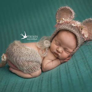 May include: A newborn baby wearing a knitted mouse hat with pink flowers and a matching knitted outfit with a tail. The baby is sleeping on a teal textured blanket.