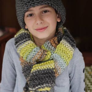Könnte beinhalten: Ein junges Kind trägt eine graue Strickmütze und einen bunten gestreiften Schal mit Quasten. Der Schal ist aus weichem, dickem Garn.