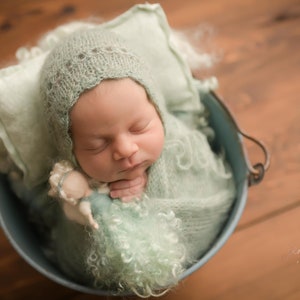 May include: A newborn baby is sleeping soundly in a green crocheted blanket and hat, nestled in a blue metal bucket. The baby is holding a small doll in their hand.