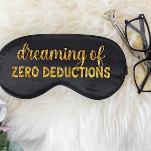May include: Black satin sleep mask with gold glitter text that reads "dreaming of ZERO DEDUCTIONS." A pair of black-framed glasses and a silver pen with a diamond-shaped top are next to the mask. The items are on a white fluffy surface.
