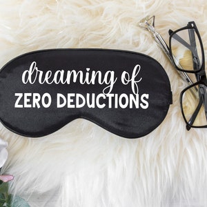 May include: Black satin sleep mask with the text "dreaming of ZERO DEDUCTIONS" in white script. The mask is next to a pair of black-framed glasses and a silver pen with a diamond-shaped top. The background is white faux fur.