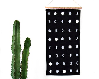 May include: Black and white moon phase tapestry hanging on a white wall. The tapestry features a repeating pattern of white moons in various phases on a black background.