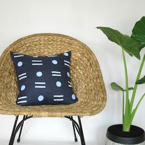 May include: A blue and white patterned throw pillow sits on a woven wicker chair. The chair has black metal legs. A green leafy plant is in a black and white striped pot in the background.
