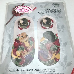 May include: A counted cross stitch kit featuring two mallard ducks with a red bow and holly. The kit includes 100% cotton floss, Aida cloth, Pellon backing fabric, a graph, a needle, and easy-to-follow instructions. The finished size of each design is 5 inches by 12 inches. The kit is from Candamar Designs Inc. and is labeled "Mallards Door Knob Decor".