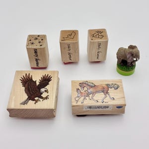 May include: A collection of five items including three small wooden stamps with floral, dolphin, and goat designs labeled Shelly Henson, a large square stamp with a bald eagle, a rectangular stamp with two horses, and a small elephant figurine.