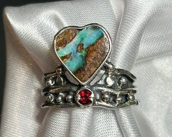 May include: A silver ring with a heart-shaped opal stone. The opal features shades of blue, green, and brown. The ring has a decorative band with a small red stone accent.