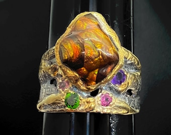 Op de afbeelding: Een unieke ring met een grote, iriserende edelsteen in oranje- en bruintinten, gezet in een gouden en zilveren band. De band is versierd met kleinere groene, roze en paarse edelstenen.