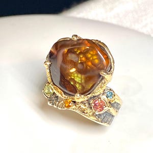 May include: A gold ring featuring a large, irregular, translucent brown fire agate as the centerpiece. The band has a textured, dark metal finish and is accented with small, round, multi-colored gemstones including yellow, pink, and blue stones.