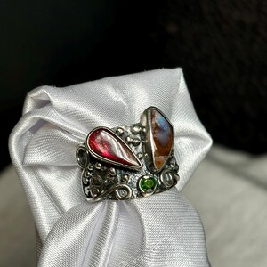 K&ouml;nnte beinhalten: Ein silberner Ring mit einem tropfenf&ouml;rmigen roten Edelstein und einem ovalen Opal mit braunen und blauen Farbt&ouml;nen. Zwischen den beiden gr&ouml;&szlig;eren Steinen ist ein kleiner gr&uuml;ner Edelstein eingesetzt. Der Ring wird auf wei&szlig;em Satin pr&auml;sentiert.