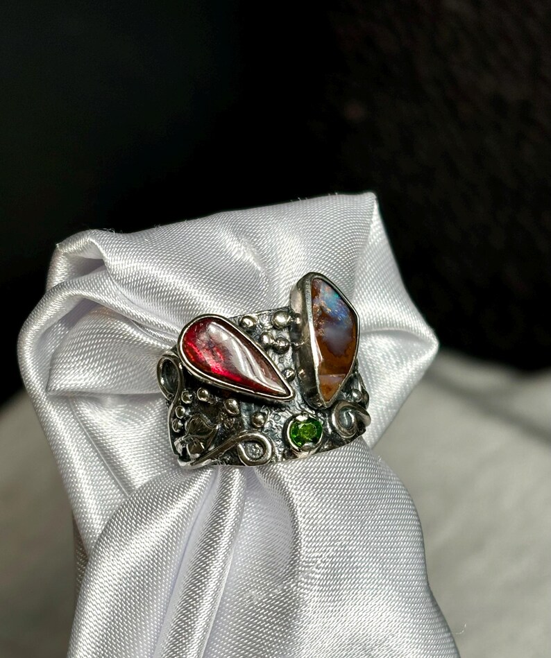 K&ouml;nnte beinhalten: Ein silberner Ring mit einem tropfenf&ouml;rmigen roten Edelstein und einem ovalen Opal mit braunen und blauen Farbt&ouml;nen. Zwischen den beiden gr&ouml;&szlig;eren Steinen ist ein kleiner gr&uuml;ner Edelstein eingesetzt. Der Ring wird auf wei&szlig;em Satin pr&auml;sentiert.