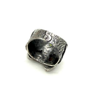 K&ouml;nnte beinhalten: Ein strukturierter Silberring mit einem einzigartigen Design. Der Ring hat eine detaillierte, raue Oberfl&auml;che und ein kleines, kreisf&ouml;rmiges Emblem. Im Inneren des Rings ist ein kleiner gr&uuml;ner Edelstein zu sehen. Der Ring ist ein Schmuckst&uuml;ck.