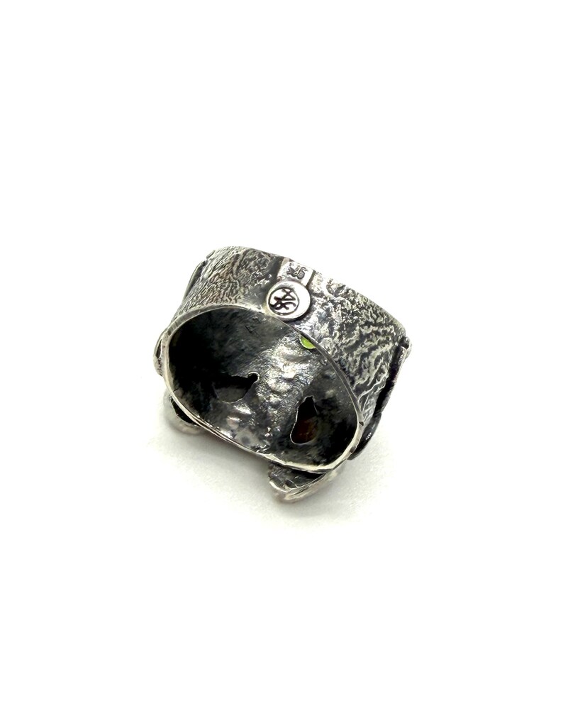 K&ouml;nnte beinhalten: Ein strukturierter Silberring mit einem einzigartigen Design. Der Ring hat eine detaillierte, raue Oberfl&auml;che und ein kleines, kreisf&ouml;rmiges Emblem. Im Inneren des Rings ist ein kleiner gr&uuml;ner Edelstein zu sehen. Der Ring ist ein Schmuckst&uuml;ck.