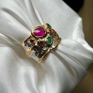 May include: A gold and silver ring with a large oval ruby center stone, surrounded by smaller emeralds and rubies. The ring has a textured silver band and a gold rim around the gemstones. The ring is displayed on a white satin fabric.