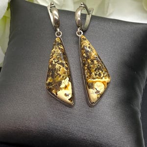 May include: A pair of silver dangle earrings with triangular amber-colored stones. The earrings feature a mix of brown, yellow, and cream tones within the amber, set in a silver frame. The earrings are displayed on a black surface.
