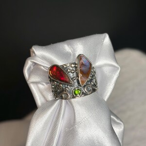 K&ouml;nnte beinhalten: Ein Silberring mit zwei gro&szlig;en, farbigen Edelsteinen und einem kleinen gr&uuml;nen Stein. Der Ring hat ein strukturiertes Band mit aufwendigen Details. Ein Edelstein ist tiefrot, der andere ist eine Mischung aus Braun, Orange und Blau.