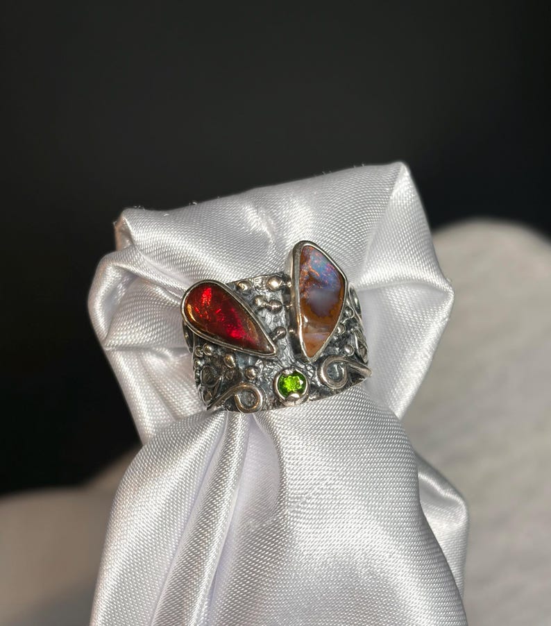 K&ouml;nnte beinhalten: Ein Silberring mit zwei gro&szlig;en, farbigen Edelsteinen und einem kleinen gr&uuml;nen Stein. Der Ring hat ein strukturiertes Band mit aufwendigen Details. Ein Edelstein ist tiefrot, der andere ist eine Mischung aus Braun, Orange und Blau.