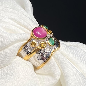 May include: A silver and gold ring with a pink ruby and green emeralds set in a textured band.